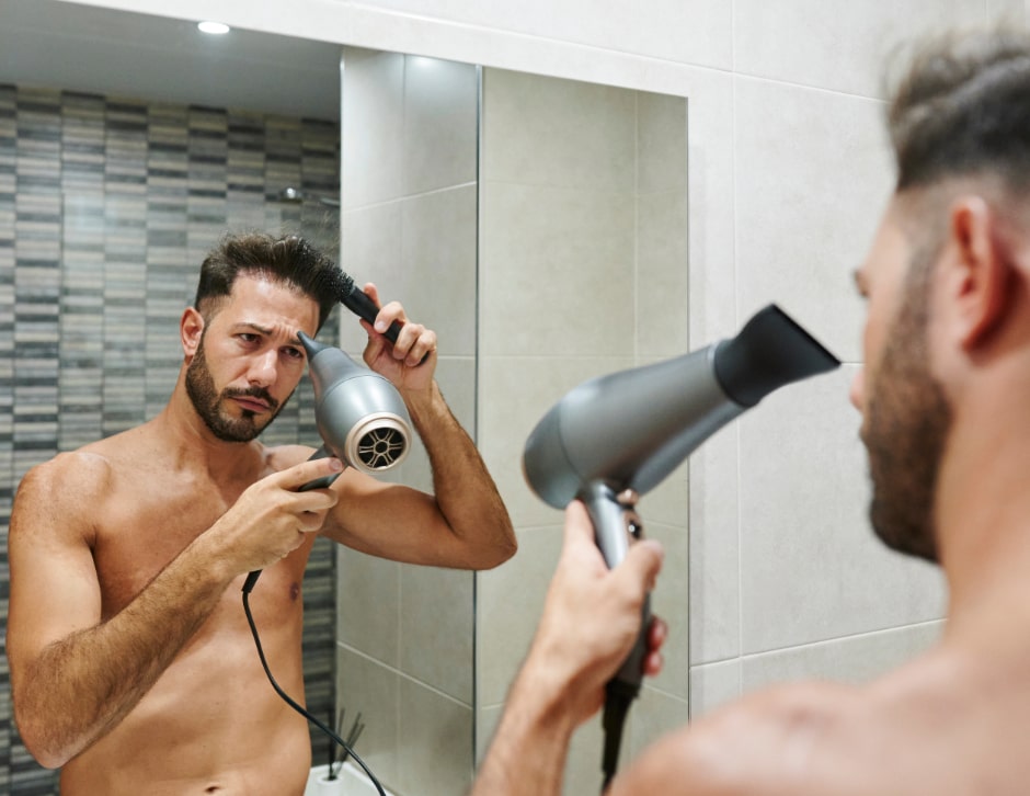 A man blow-drying and combing his hair back while looking in the mirror, revealing some hair loss.