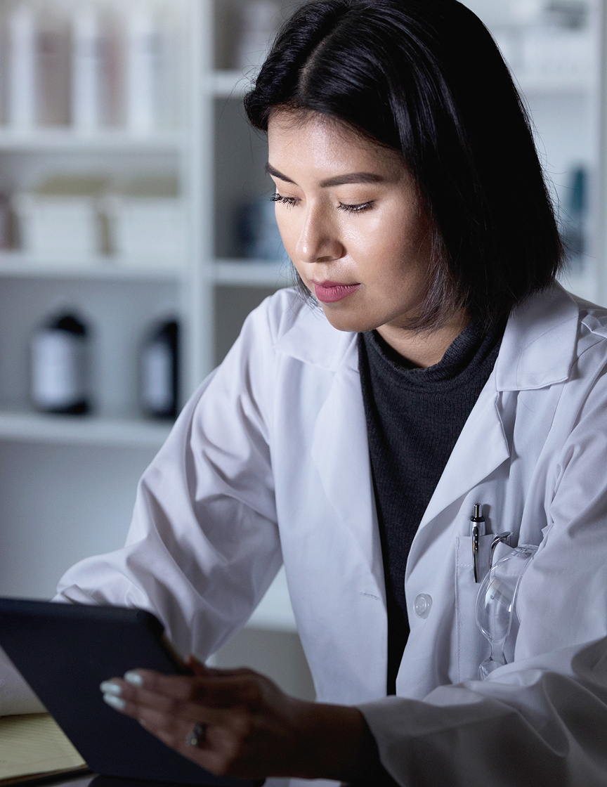 Female pharmacist in a lab coat reviewing information on a tablet, with pharmacy shelves and bottles in the background