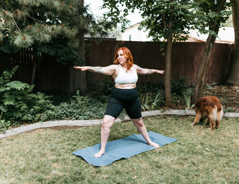 A wide shot of a woman doing yoga in her backyard, with her dog comfortably grazing at her side.