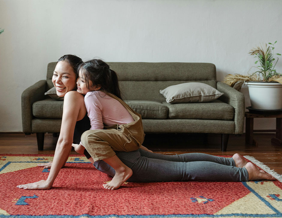 A woman balancing her daughter on her back as she does yoga, both smiling in the sunshine from the living room window.