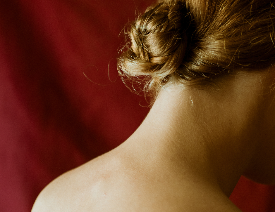 Close-up of a woman's neck and shoulder with braided hair, seen from behind against a deep red backdrop