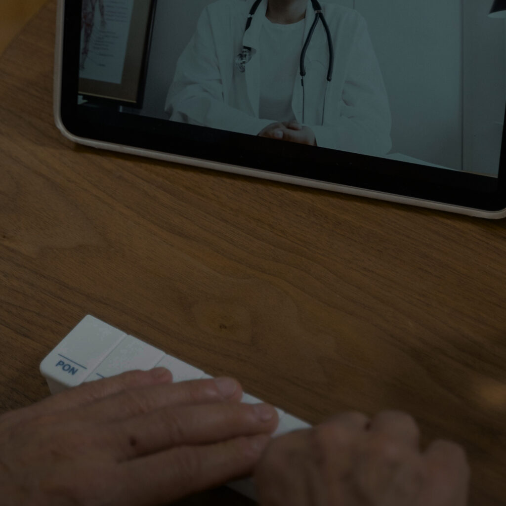 Person's hands holding a pill organizer on a wooden table during a video call with a doctor shown on a tablet screen