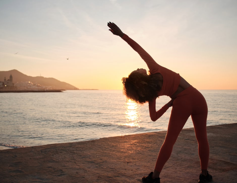 Woman doing yoga stretches on a sandy beach while the sun sets over the horizon in front of her.