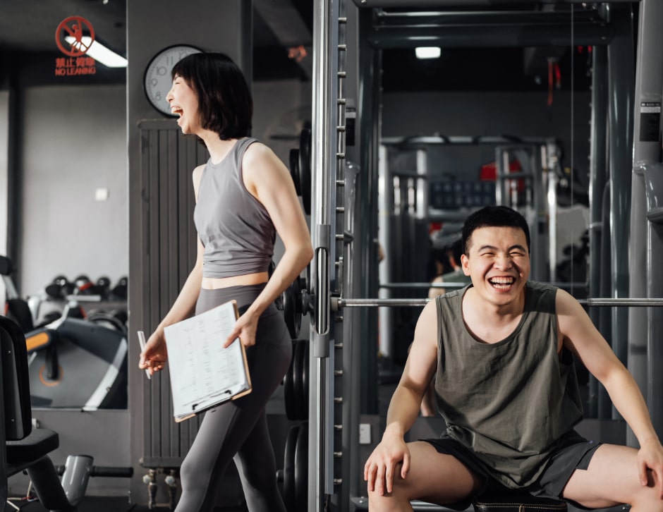 A man and woman laugh around a bench press after finishing a workout.